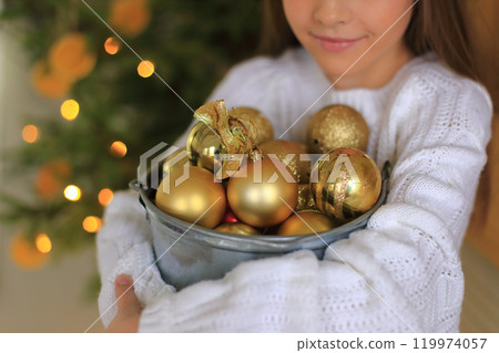 a pair of hands holding an array of shimmering golden ornaments, perfect for decorating a Christmas tree. The warm glow of twinkling lights and festive decorations in the background enhances the a pair of hands holding an array of shimmering golden ornaments, perfect for decorating a Christmas tree. The warm glow of twinkling lights and festive decorations in the background enhances the 119974057