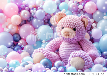 A crocheted handmade bear toy in pastel light violet color sitting among colorful purple pink teal balls and air balloons. Birthday card celebration 119974154