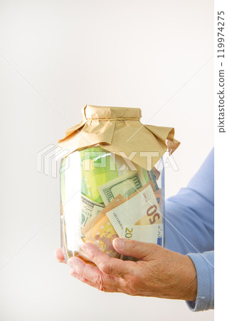 A persons hands are holding a glass jar filled with paper money. The jar is covered with a brown paper bag A persons hands are holding a glass jar filled with paper money. The jar is covered with a brown paper bag 119974275