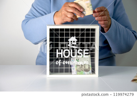 A person in a blue sweater is putting money into a white box with a black grid and the words House of our dreams on it 119974279