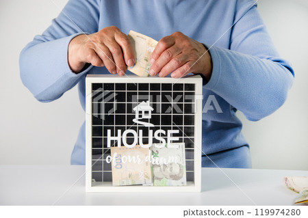 A person puts money into a white money box with a black background and a house drawn on it 119974280