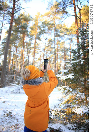A young man stands in a winter forest, capturing the beauty of the snowy landscape with his smartphone. A young man stands in a winter forest, capturing the beauty of the snowy landscape with his smartphone. 119974781