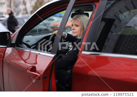 A professional businesswoman sits confidently in her stylish red car, parked outside a modern business center.  119975521