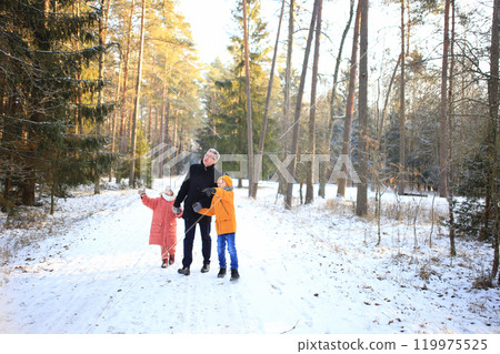 A father enjoys a leisurely walk with his two children through a beautiful snowy forest. The scene captures the joy of family time amidst the serene winter landscape, with snow-covered trees creating 119975525