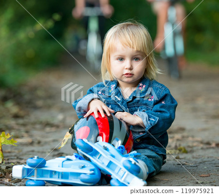 Cute little girl on roller skates sitting on road in the park Cute little girl on roller skates sitting on road in the park 119975921