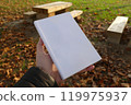 A white book held in a hand against a backdrop of wooden benches and autumn leaves 119975937