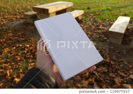 A white book held in a hand against a backdrop of wooden benches and autumn leaves 119975937
