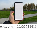 A hand holding a smartphone with a blank white screen against a backdrop of a park with green grass and trees. 119975938