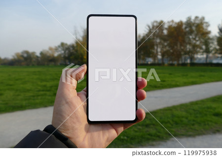 A hand holding a smartphone with a blank white screen against a backdrop of a park with green grass and trees. 119975938