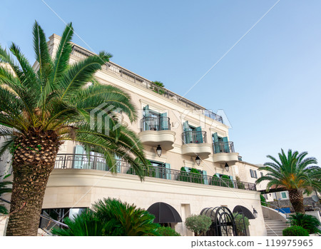 Elegant Mediterranean-style hotel with balconies and palm trees under a clear blue sky, perfect for a luxurious getaway Elegant Mediterranean-style hotel with balconies and palm trees under a clear blue sky, perfect for a luxurious getaway 119975965
