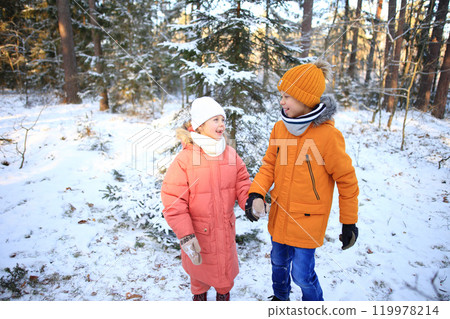 A brother and sister joyfully walk together through a serene snowy forest, surrounded by tall trees blanketed in white.  119978214