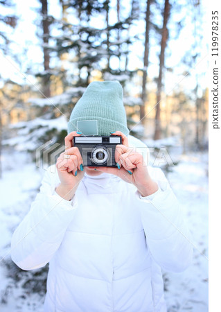 a woman holding a vintage camera, set against the stunning backdrop of a snowy winter forest. Dressed warmly for the cold, she embodies a sense of nostalgia and creativity, ready to capture the beauty 119978235