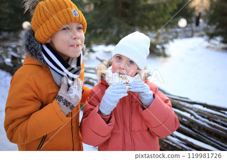 A brother and sister share a delightful moment as they eat gingerbread cookies outdoors against the backdrop of a snowy forest.  119981726