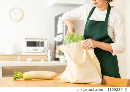 A middle-aged to senior woman taking out groceries from her own bag in the living room at home 119982943