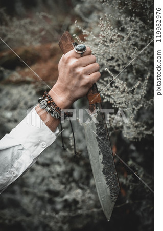 Authentic handmade knife in female hand with bracelets on atmospheric forest background in late autumn, vertical format, selective focus. 119982976
