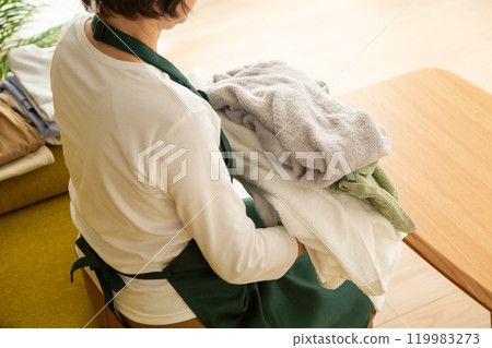 穿著圍裙的中高級女性在客廳的沙發上疊衣服 穿著圍裙的中高級女性在客廳的沙發上疊衣服 119983273