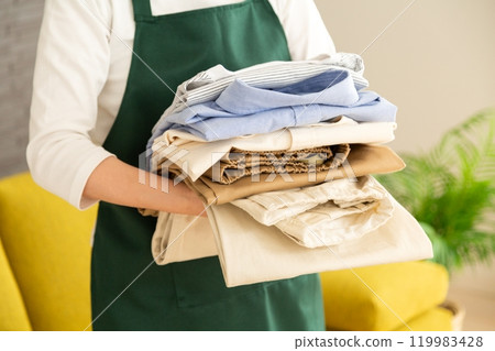 A middle-aged to senior woman wearing an apron folding laundry on the sofa in the living room A middle-aged to senior woman wearing an apron folding laundry on the sofa in the living room 119983428