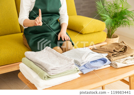 一位中年至老年婦女穿著圍裙，在客廳的沙發上疊衣服，舉起食指 119983441