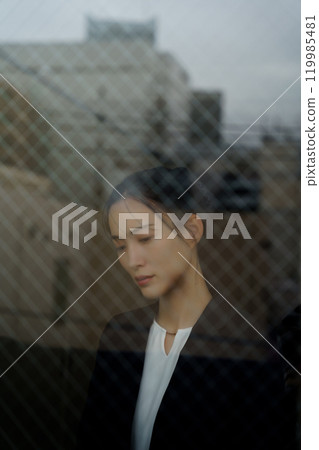 一位穿著西裝的年輕女子,表情憂鬱,透過玻璃倒映著城市景觀 一位穿著西裝的年輕女子,表情憂鬱,透過玻璃倒映著城市景觀 119985481
