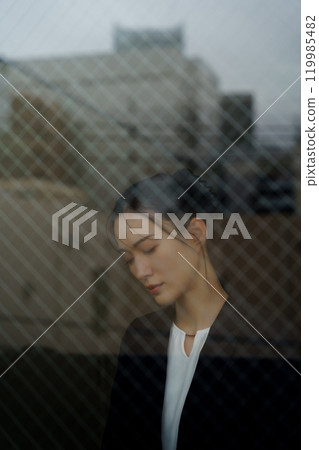 一位穿著西裝的年輕女子,表情憂鬱,透過玻璃倒映著城市景觀 一位穿著西裝的年輕女子,表情憂鬱,透過玻璃倒映著城市景觀 119985482
