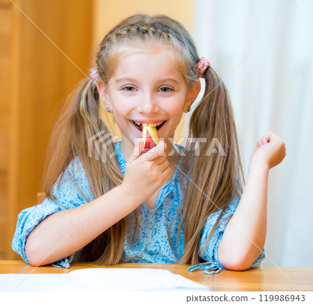 Schoolgirl eating a slice of apple at her desk 119986943