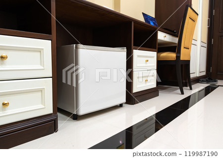 mini fridge in low angle view, inside it is empty shelves placed under wooden counter and next to wood closet and chair in a luxury hotel room. 119987190