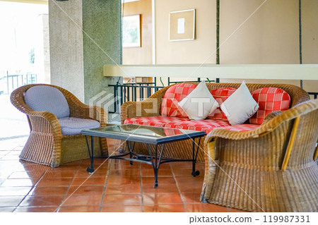 Traditional Thai rattan wicker furniture set consists of a sofa, armchairs, and a glass coffee table, placed in the reception or relaxing zone of the resort. Wall decorated with handcraft pictures. 119987331