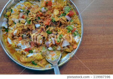 Bhel puri chaat served in a plate with a spoon, showcasing puffed rice, sev, chopped onions, and tomatoes in a vibrant mix. Traditional authentic snack food originating in Mumbai, India 119987396