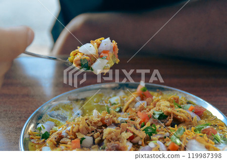 A foodie taking a spoonful of Bhel puri chaat served in a plate made of puffed rice, sev, chopped onions, and tomatoes in a vibrant mix. Traditional authentic snack food originating in Mumbai, India 119987398