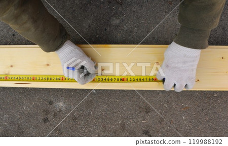 A man in work clothes and gloves uses a tape measure and pen to make markings on a light-colored wooden board lying on the asphalt 119988192