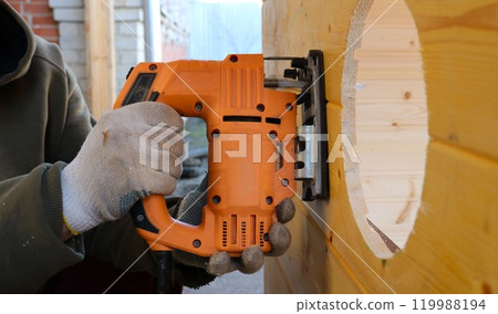 Worker in dirty work gloves cutting a circle in a wooden wall with an electric jigsaw, side view, jigsaw work vertically to make round holes in a wooden wall of a farm building 119988194