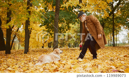 Elderly man and his dog on cozy walk surrounded by golden autumn leaves, enjoying cool breeze and playing together in fall park. Elderly man and his dog on cozy walk surrounded by golden autumn leaves, enjoying cool breeze and playing together in fall park. 119989073