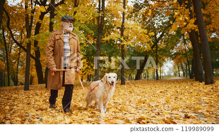 Elderly man, dressed stylish clothes in warm coat walking with his cheerful dog through golden hues of autumn park. 119989099