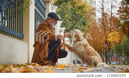 Elderly, stylish dressed man playing, training his purebred dog, Golden retriever at park in autumn. Walking outdoor. Elderly, stylish dressed man playing, training his purebred dog, Golden retriever at park in autumn. Walking outdoor. 119989195