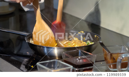 A cook stirs fry Pad Thai noodles in a pan with spatulas on electric stoves. Sauce dishes for cooking traditional Pad Thai. A cook stirs fry Pad Thai noodles in a pan with spatulas on electric stoves. Sauce dishes for cooking traditional Pad Thai. 119989339