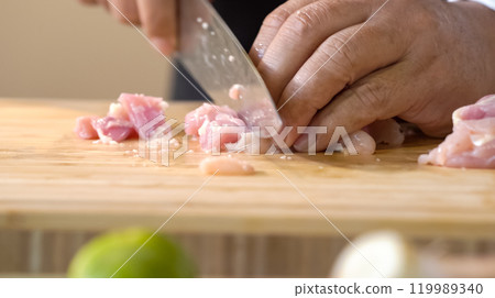 A chef using a kitchen knife slices uncooked chicken meat on a chopping board, the close up view of preparing food ingredients. 119989340
