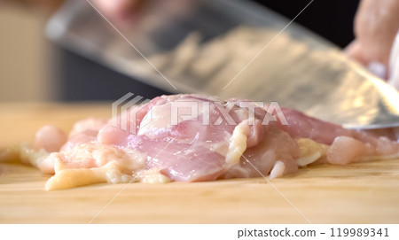 A chef using a kitchen knife slices uncooked chicken meat on a chopping board, the close up view of preparing food ingredients. A chef using a kitchen knife slices uncooked chicken meat on a chopping board, the close up view of preparing food ingredients. 119989341