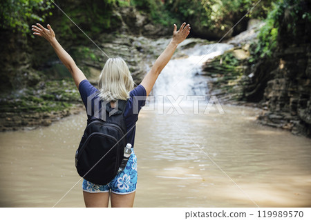 Female traveler looking at waterfall and raising her hands up Female traveler looking at waterfall and raising her hands up 119989570