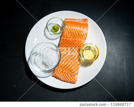 Flatlay Toptable raw Salmon or trout steak fillet on white dish with salt, oil, wasabi and micro herb in studio light on dark background. Clipping Paths. Flatlay Toptable raw Salmon or trout steak fillet on white dish with salt, oil, wasabi and micro herb in studio light on dark background. Clipping Paths. 119989717