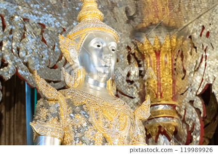Deity statue standing straight made of silver, wearing silver gold costume with Chada on top of his head. Surrounded by sparkleling decoration. Deity statue standing straight made of silver, wearing silver gold costume with Chada on top of his head. Surrounded by sparkleling decoration. 119989926
