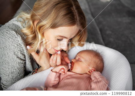 Beautiful mother gazes with love at sleeping newborn baby, dressed in pink onesie and has pacifier in mouth. Serene moment at home, reflects deep bond, tenderness, and affection between mom and child 119990812