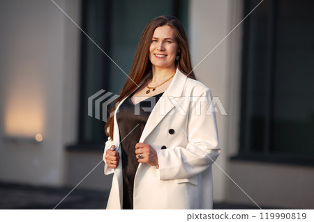 Portrait Plus size smiling business woman stands outdoors in urban setting at city, wearing fashionable black dress with white double breasted blazer outfit. Model with long brown hair 119990819