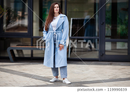 Plus size young woman stands outdoors modern urban setting contemporary city, wearing stylish light blue trench coat, white t-shirt and blue jeans, with long brown hair and wears casual white sneakers Plus size young woman stands outdoors modern urban setting contemporary city, wearing stylish light blue trench coat, white t-shirt and blue jeans, with long brown hair and wears casual white sneakers 119990839