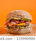 Close-up of a cheeseburger with bacon, pickles, and sauce on a wooden surface against an orange background. Studio food photography. American fast food and snack concept. Design for poster, banner 119990900