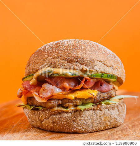 Close-up of a cheeseburger with bacon, pickles, and sauce on a wooden surface against an orange background. Studio food photography. American fast food and snack concept. Design for poster, banner Close-up of a cheeseburger with bacon, pickles, and sauce on a wooden surface against an orange background. Studio food photography. American fast food and snack concept. Design for poster, banner 119990900