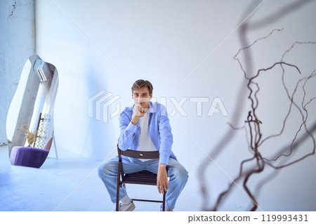 portrait of a handsome young adult man in a blue shirt sitting on a chair on a blue background 119993431