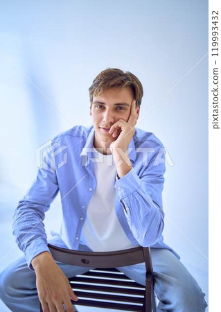 portrait of a handsome young adult man in a blue shirt sitting on a chair on a blue background portrait of a handsome young adult man in a blue shirt sitting on a chair on a blue background 119993432