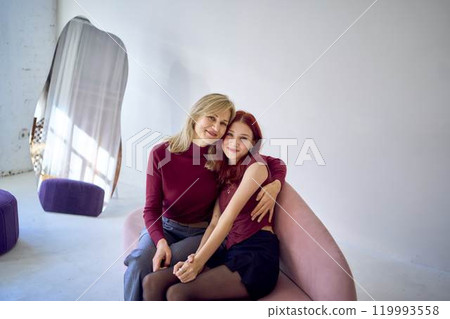 attractive middle-aged mother and teenage daughter on a pink sofa in a spacious white studio 119993558