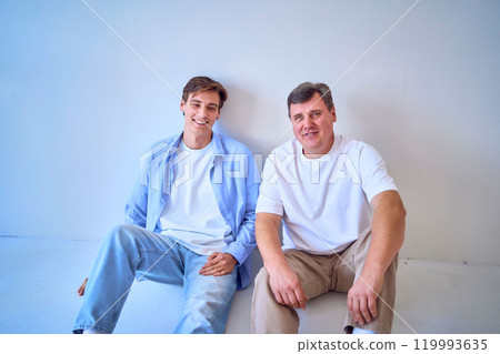 a portrait of a middle-aged man and his adult son sitting by the wall on a white background a portrait of a middle-aged man and his adult son sitting by the wall on a white background 119993635