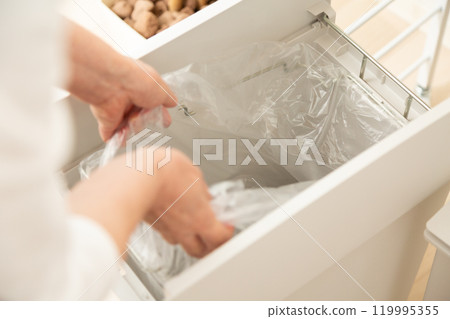 Hands of a middle-aged to senior woman putting a garbage bag into a kitchen trash can 119995355